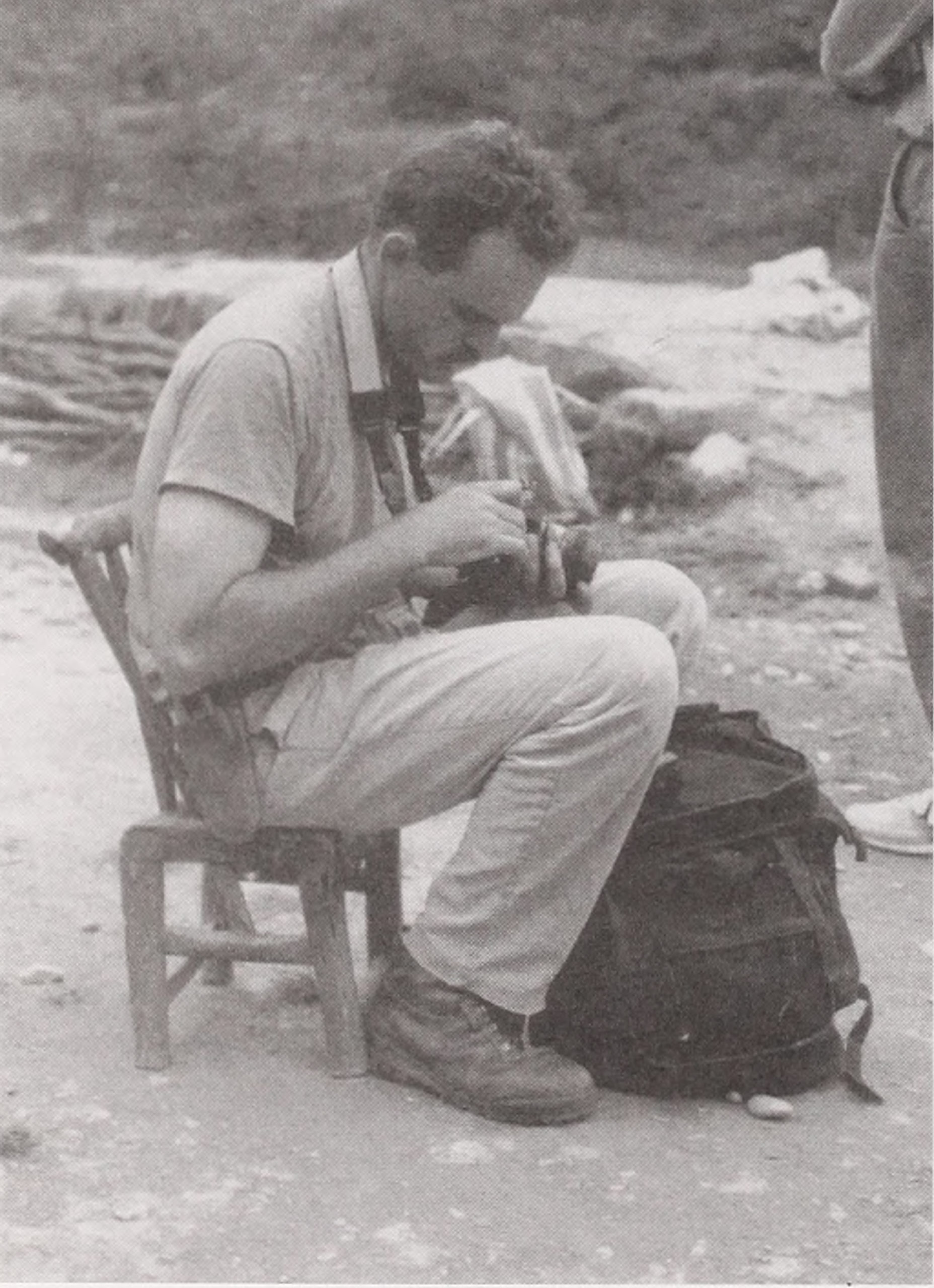 Black-and-white photograph of man sitting on small chair with camera on lap