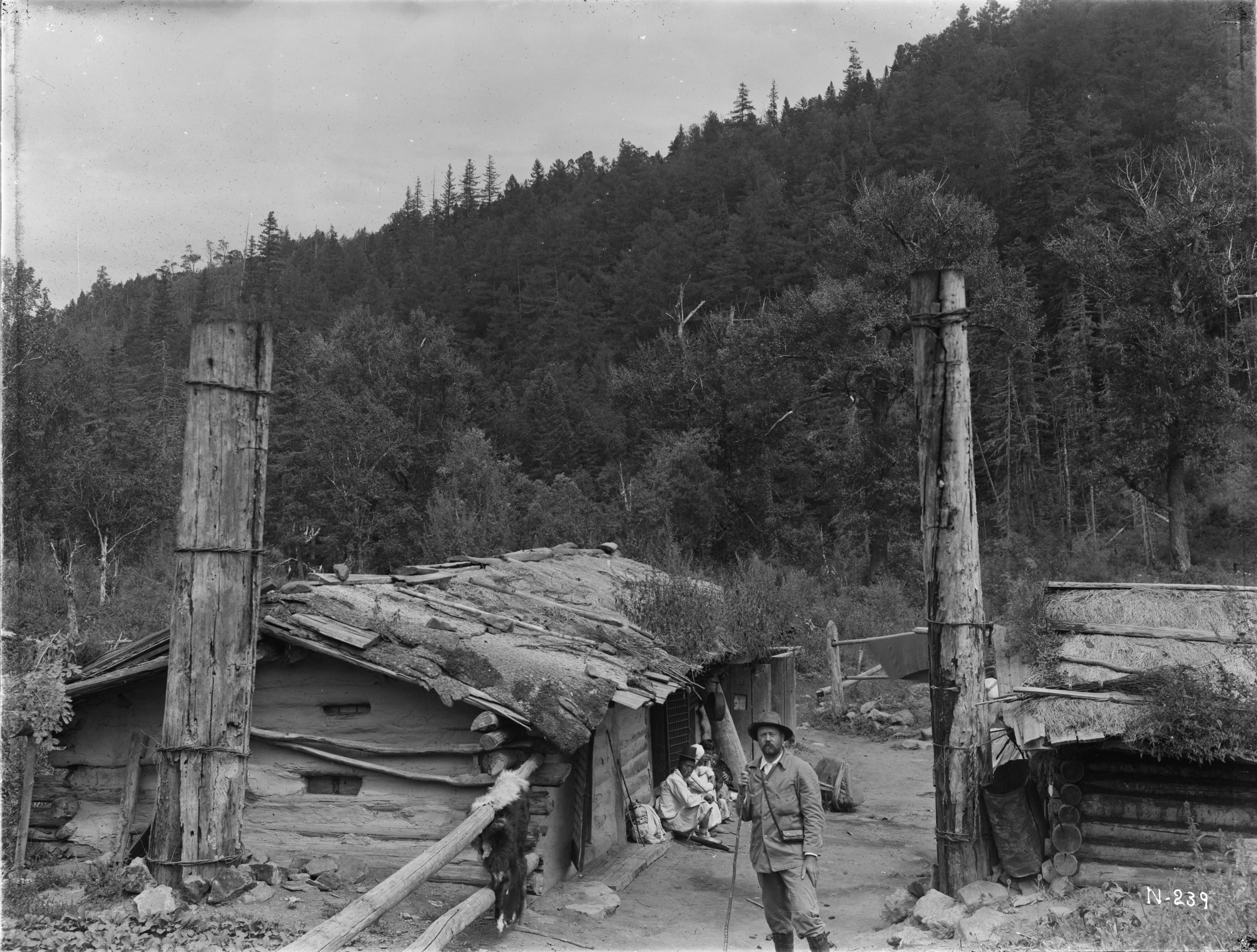 Ernest Wilson stands before an inn at Tokuitsudo on the Tumen-Yalu divide, North Kankyo Province in August 1917. A mixed forest of Korean poplar (Populus suaveolens) and Khingan fir (Abies nephrolepis) grow on the hill behind.