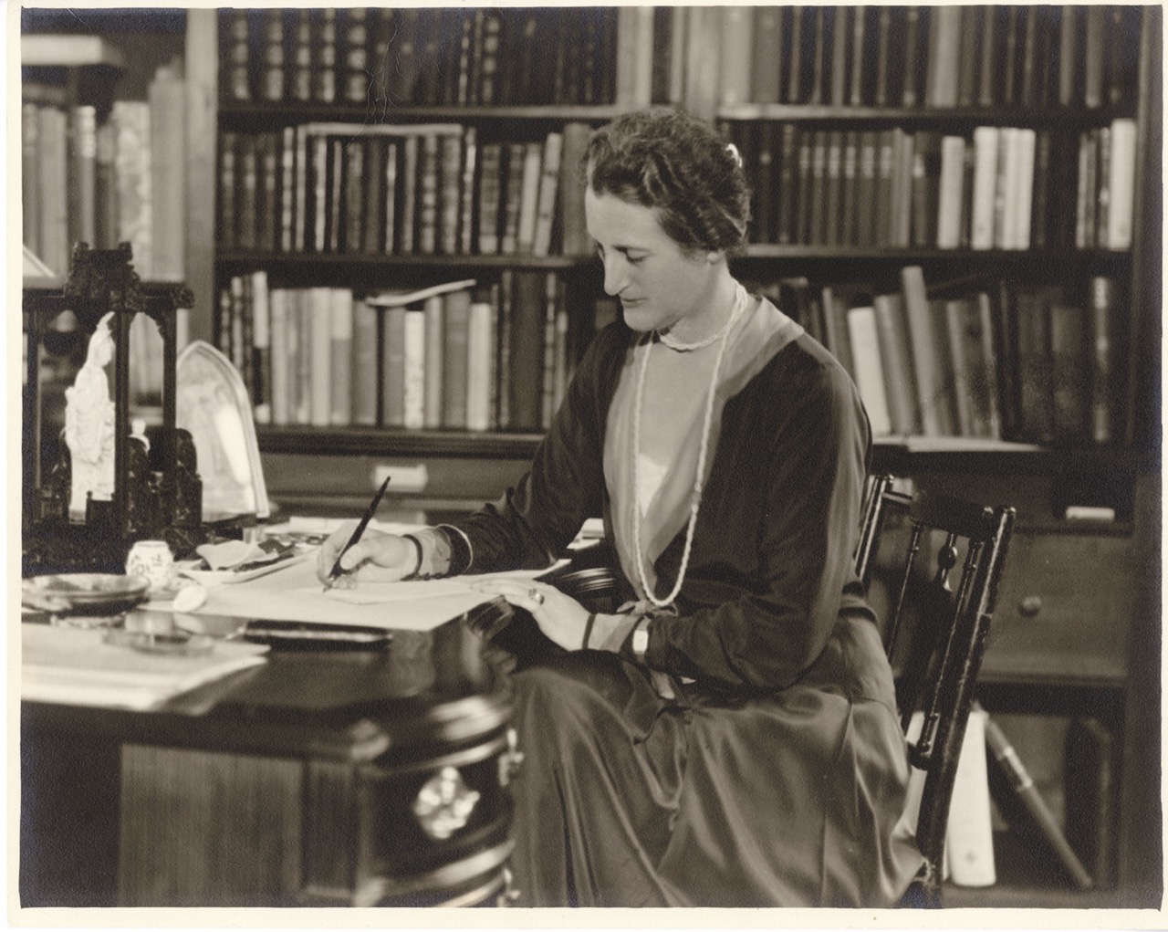 Black and white photo of woman seated writing at table with books in the background.