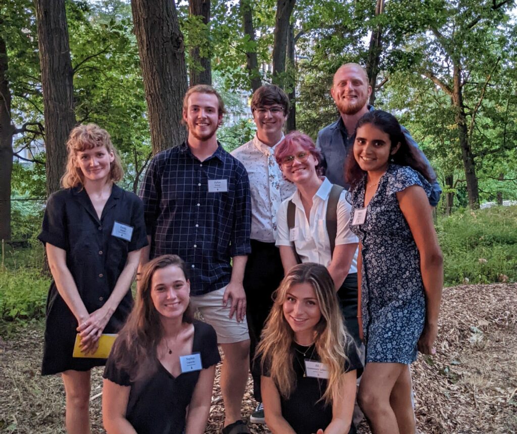 A group of eight people, some standing and some kneeling, pose and smile for a photo outdoors in a wooded area, all wearing name tags and casual summer clothing.
