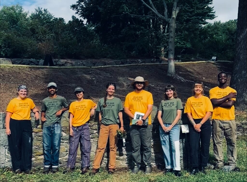 Eight people stand outdoors in a row, some wearing yellow shirts and others in green shirts, all smiling. Trees and a grassy area are visible in the background.