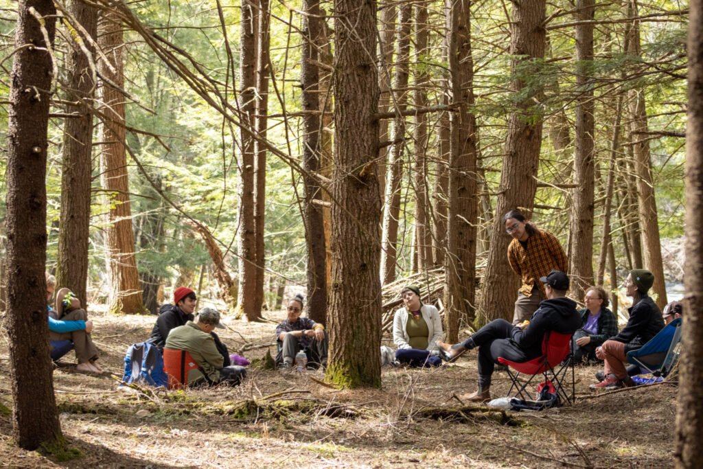 A group of people sit in a circle on chairs and logs in a forest, listening to a person standing and speaking among tall trees, with sunlight filtering through the branches.