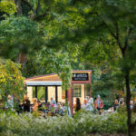 Several people mill about amongst the trees and a Mobile Visitor Center