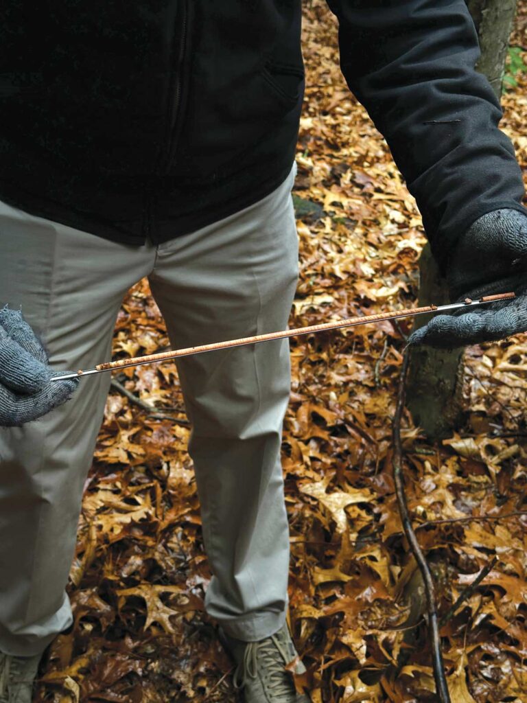 Person standing on leaf-covered ground, holding a thin metal rod containing a pencil-thick piece of wood.