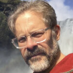 headshot of matthew battles with beard and clear-rimmed glasses wearing a red sweatshirt with a waterfall visible int h background