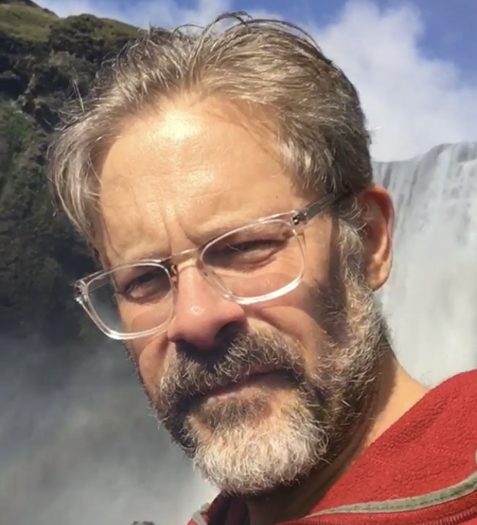 headshot of matthew battles with beard and clear-rimmed glasses wearing a red sweatshirt with a waterfall visible int h background
