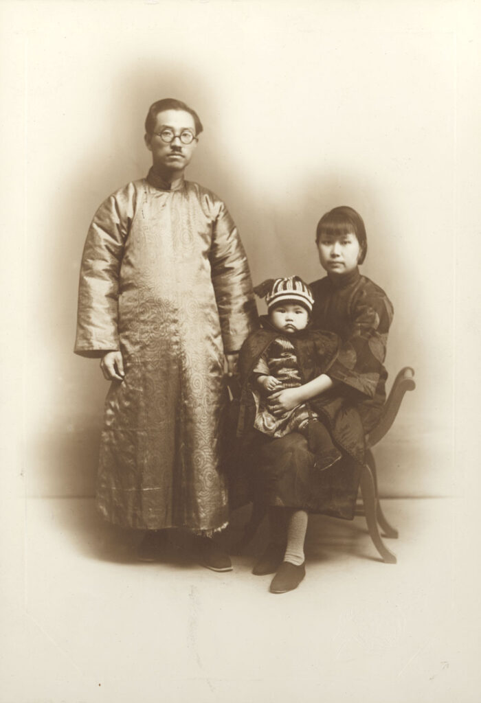 Black and white photograph of man standing, woman seated with a small child on her lap