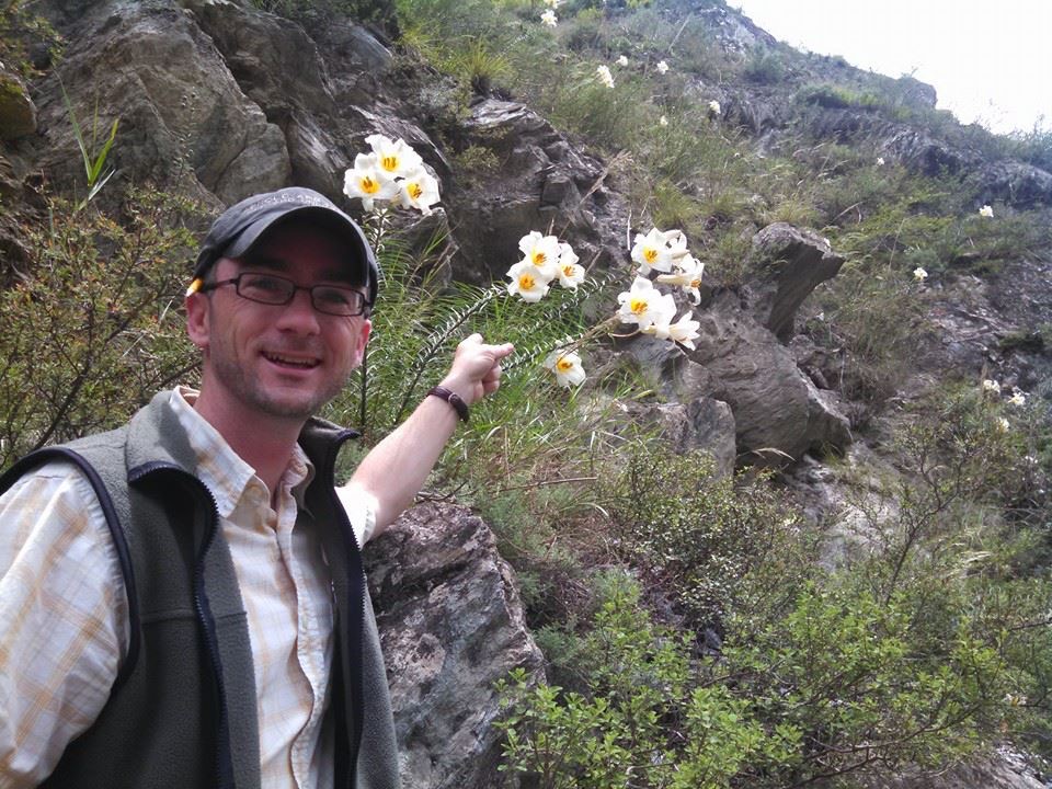 A man points to a white flower on a mountainside