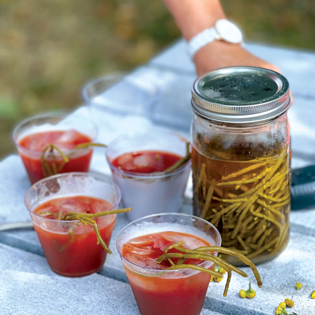a jar filled with sea beans and with a red-colored liquid, along with four cups of the beverage