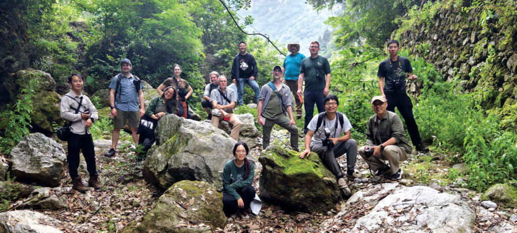 14 people posing for a photo in a wooded setting, some are standing, some are crouching or sitting on large rocks in the landscape.