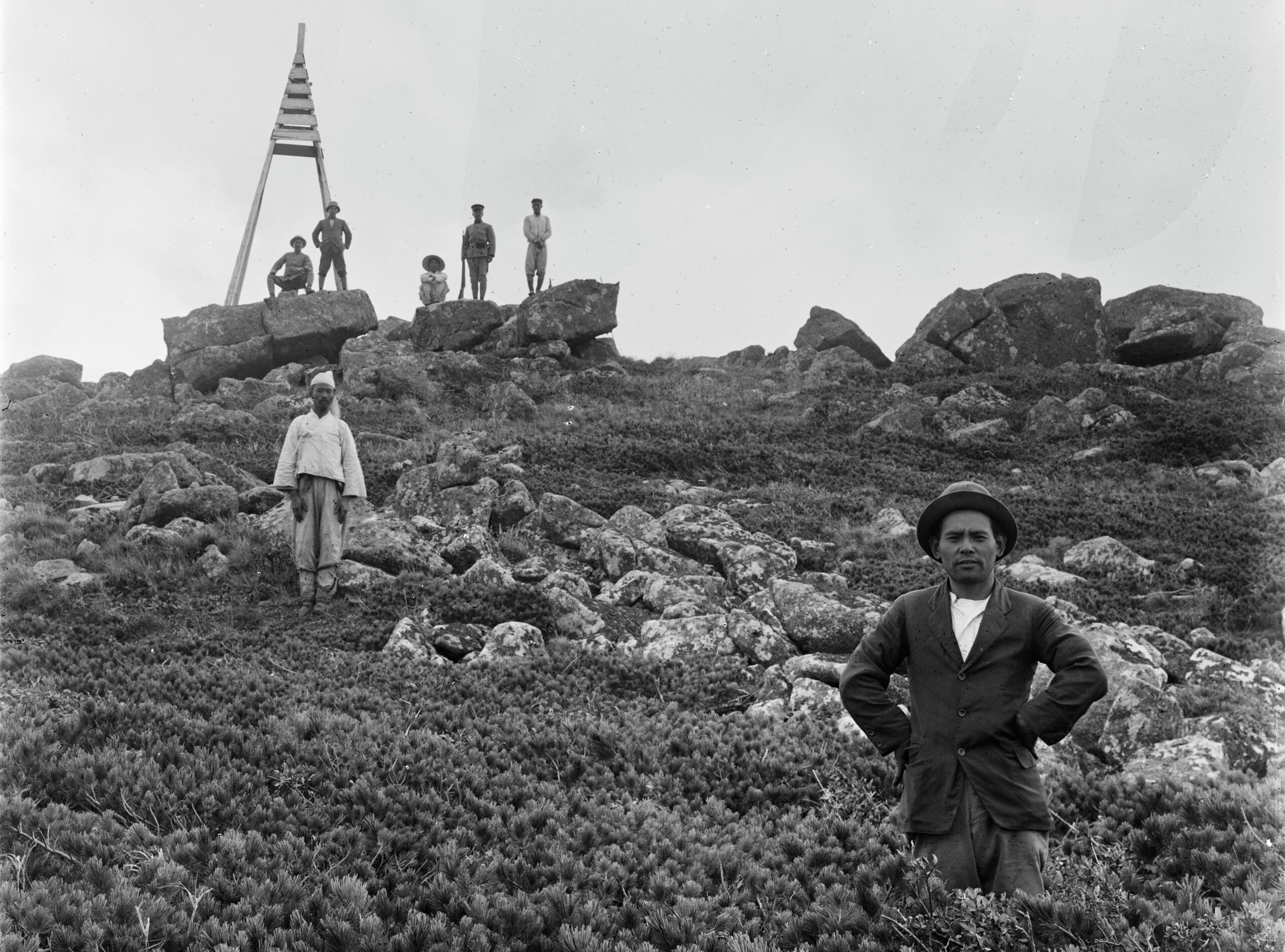 Wilson's personal assistant Mr. Morita stands in a growth of Japanese stone pine, a Korean man stands further up the slope by a pile of boulders, in the distance, at the summit of the mountain an A-frame wooden structure stands, just in front of it on large boulders, from left to right, is a seated man, beside him is Mr. S. Tukodo, then a seated Korean man, beside him a man in uniform with a rifle, and another Korean man